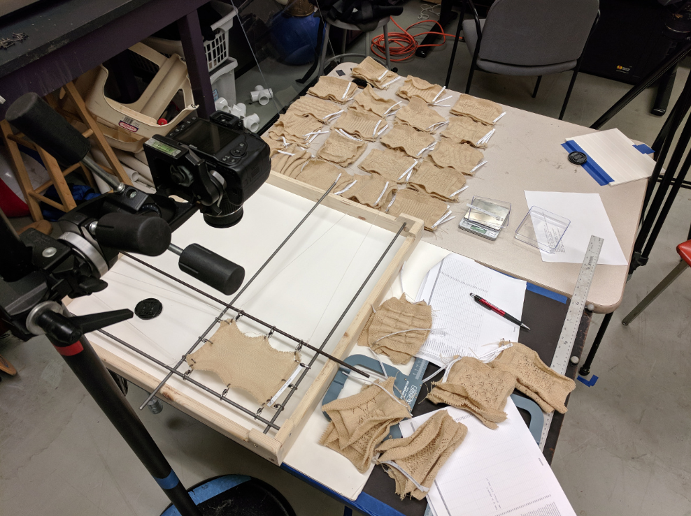 A photograph of the table with our data measurement setup, along with piles of patches that are about to be measured and have recently been measured. One patch is attached to the rods and clips used for stretching.